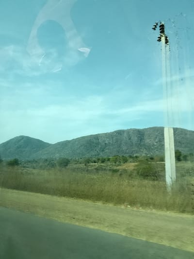This image depicts an outdoor landscape scene in Jibam, Nigeria. The foreground features a dark, paved road extending diagonally from the lower left towards the center, alongside a strip of light brown, dry earth. The midground consists of extensive fields of dry, yellowish-brown grasses and scrubland, dotted with scattered green trees and bushes. In the background, two prominent, dark green, tree-covered hills or mountains rise against a clear, light blue sky with minimal cloud cover. A tall, slender, light-colored utility pole is positioned on the far right, topped with dark utility components. Reflections, including an inverted U-shape and irregular light patterns, are visible across the transparent surface through which the photograph was taken, predominantly in the upper left, along with vertical streaks on the right side. No individuals, animals, or discernible actions are present within the scene.