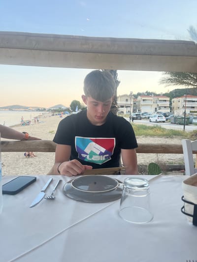 A young male with dark hair, wearing a black t-shirt with a colorful geometric logo, is seated at a table covered with a white tablecloth. He is looking down at a piece of paper in his hands. On the table in front of him are a grey plate, a clear glass, and a set of silverware including a knife and a fork. A black smartphone is also visible on the table.
The scene is set outdoors, possibly a beachside restaurant, indicated by the sandy beach and the ocean in the background. Several people are visible on the beach, some sunbathing, others walking. In the distance, there are buildings and trees. The lighting suggests it is late afternoon or early evening, with a warm, soft glow in the sky. The overall atmosphere appears relaxed and casual.
A sign on a building in the background seems to have Greek writing, possibly indicating a parking area. The overall location context is Nea Peramos, Greece.