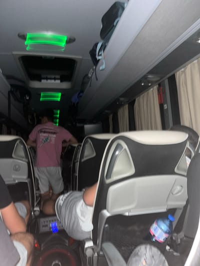 The image shows the interior of a bus. Seats are visible, occupied by several individuals. One person is wearing a pink shirt and light-colored pants. There is a water bottle in the seat pocket of one of the chairs. Green lights illuminate a section of the wall. The context of the location being in Lesichovo, Bulgaria is not discernible from the image itself.