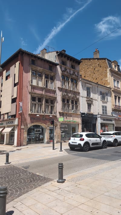 The image captures a street scene in Villefranche-sur-Saône, France, under a clear blue sky with a wispy cloud formation. The foreground shows a paved street and sidewalk with several dark grey bollards. The sidewalk is a mosaic of large light-colored square tiles and a cobblestone area. In the midground, a row of buildings lines the street.
The building on the left, with a reddish-pink facade, features a prominent sign above an arched storefront that reads "PHOTO G. PICHON". The storefront itself displays photographs and mannequins. A smaller sign on the left facade reads "SERVICE G. PIC HON". Next to it, a sign displays the FUJI logo and text.
Adjacent to the "PHOTO G. PICHON" building is another building with ornate stone carvings around its upper windows. Further down the street, a building with a textured, tan-colored stone facade is visible, followed by a lighter-colored building with more windows. Storefronts line the ground floor of these buildings. One storefront has a sign with the word "PRESSING" and "PRESTIGE". Another sign indicates "COUETTE Housse Canapé Rénovation Retouche". Further along, a store is labeled "CALZEDONIA".
Two white cars are parked on the street. The car in the foreground is a Citroën C3 with the license plate "GY 395 AZ". A second white car, possibly an SUV, is parked behind it. A bicycle is visible through the window of the "COUETTE" shop. A person in a light orange shirt is seen standing in front of the "CALZEDONIA" store. Traffic lights are visible above the "PHOTO G. PICHON" storefront. Antennas and a chimney are present on the rooftops.