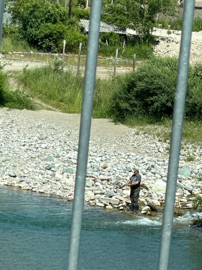 A man is fishing in a river with a rocky shore. He is wearing waders and a hat. The river is flowing rapidly, and there are trees and bushes in the background. The scene is viewed through metal bars, suggesting it may be taken from a bridge or a similar structure. The location is in the city of Dulovine, Montenegro.