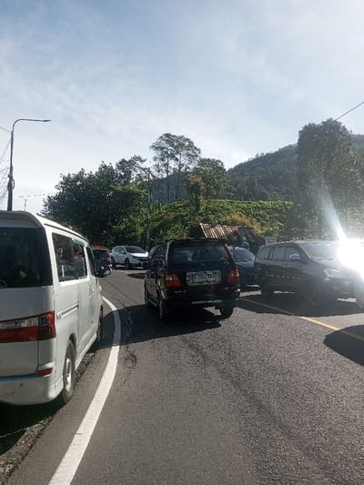 The image displays an outdoor scene of a vehicular queue on an asphalt road in Ka Thaung (Upper), Myanmar, under a bright, partly cloudy sky. On the left side of the frame, a light-colored minivan is stationary on the road's shoulder, marked by a solid white line. In the adjacent lane, a dark-colored SUV with an Indonesian license plate "B 3344 KVB" is stopped, facing away from the camera. Behind it, a red motorcycle and a white SUV are partially visible, with further vehicles extending into the background, suggesting traffic. To the right, another dark SUV is partially obscured by intense lens flare originating from the right side of the frame. The road surface shows signs of wear and is bordered by a solid yellow line on the right. A tall street light pole with multiple wires is positioned on the left. The background features a large, densely vegetated mountain or hill, covered with numerous trees and green foliage. A dilapidated structure with a rusty corrugated metal roof is visible on the right side of the road, at the base of the vegetated slope. No individuals are distinctly visible.