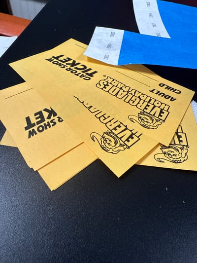The image shows several yellow tickets and a couple of blue wristbands on a dark surface. The tickets are for "Everglades Holiday Park" in Fort Lauderdale, United States. They are marked "Gator Show Ticket" and have spaces for "Adult" and "Child" designation. The blue wristbands have numbers printed on them.