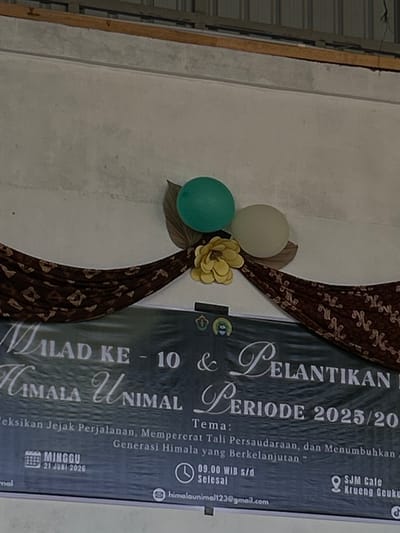The image shows a banner displayed on a wall, likely for an event. The banner has text and decorative elements. At the top of the image, a portion of a ceiling structure is visible, with what appears to be a metal roof and wooden beams. Below this, a textured wall serves as the backdrop for the banner. The banner itself is dark-colored with white and light-colored text. Prominently displayed are the words "MILAD KE - 10 & PELANTIKAN" and "HIMALA UNIMAL PERIODE 2025/2026". There is also a section for "Tema:" followed by text in a smaller font. On the left side of the banner, a calendar icon shows "MINGGU 21 JUNI 2026". On the right side, a clock icon indicates "09.00 WIB s/d Selesai". Further details on the banner include a Gmail address and a location that reads "SJM Cafe Krueng Geukuh". Two balloons, one teal and one off-white, are attached to the wall above the banner, along with a yellow paper flower and some brown leaf decorations. The overall setting appears to be an indoor event space. There are no people visible in the image. The city of Palohlada, Indonesia is not mentioned or depicted in the image.