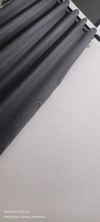The image shows a close-up of dark gray curtains with visible folds and texture, hanging from a rod. The curtains are angled diagonally across the frame. To the right of the curtains is a plain, off-white wall. The lower left corner of the image displays a timestamp and location: "2026/06/16 23:49" and "Kecamatan Ciomas, Indonesia". There are no people or other objects visible in the scene. The lighting is dim, casting subtle shadows on the curtains.