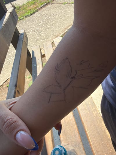 The image shows a person's forearm with a rough sketch of a flower or butterfly drawn on it. The person's hand is visible in the foreground with manicured nails, featuring a French tip design with blue accents. They are seated on a wooden bench, with the background showing gravel and some green vegetation, suggesting an outdoor setting. A plastic water bottle is partially visible at the bottom of the frame. The overall lighting indicates it is daytime. No information about Dinkata, Bulgaria, is present in the image.