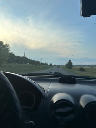 The image captures a perspective from inside a car, looking forward through the windshield towards a rural road. The car interior is visible in the foreground, with the steering wheel on the left and the dashboard in the center and right. A dashcam is mounted at the top center of the windshield. The road ahead stretches into the distance, flanked by fields and trees. Utility poles line the side of the road, indicating a developed, albeit rural, area. The sky is partly cloudy, with a soft, diffused light suggesting either early morning or late afternoon. There are no people visible in the image. The overall scene depicts a journey through the Moldovan countryside, potentially near Carabetovca, Moldova, based on the information provided.