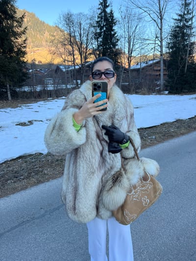 An adult female is depicted standing on a paved road surface in Saanen, Switzerland, during daylight hours. She is wearing a voluminous, light-toned fur coat, a neon green long-sleeved top, white wide-leg pants, and dark rectangular sunglasses. Her left hand holds a dark-colored smartphone with a blue textured grip, positioned as if taking a selfie. Her right hand, gloved in black leather with a visible green buckle, holds a small black object resembling a camera or lens. A tan suede handbag with white embroidery and fur accents hangs from her right arm. She is smiling, revealing teeth.
The immediate environment includes patches of snow and bare ground with dry vegetation bordering the paved road to her left. The background features a mountainous landscape characteristic of the Swiss Alps. A prominent green mountain slope, partially illuminated by sunlight revealing yellowish-brown sections, dominates the left background. Numerous bare deciduous trees and dark coniferous trees are scattered across the mid-ground and background. Several traditional wooden chalets with dark roofs are visible nestled among the trees and at the base of the mountains. The sky is clear and blue.