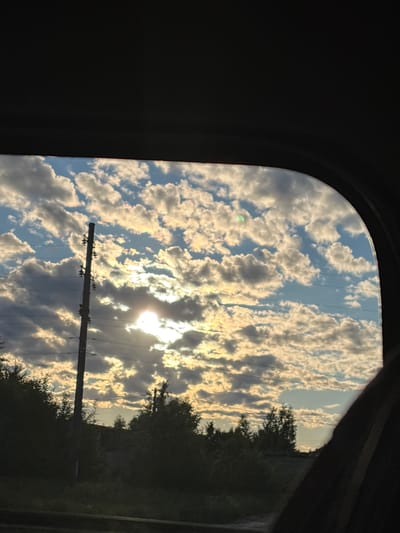 The image shows a view of the sky and trees through a vehicle window. The sky is filled with cumulus clouds illuminated by the setting sun, which is partially obscured by the clouds. A telephone pole with insulators and wires stands vertically in the left portion of the frame. The foreground is dark, suggesting the interior of a vehicle. Trees and foliage are visible at the bottom of the frame, partially silhouetted against the sky. The scene appears to be a rural or suburban landscape, potentially on the outskirts of Makaryev, Russia, given the presence of the telephone pole and the natural surroundings.