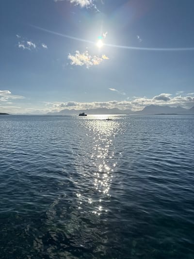This image shows a wide view of the sea, with a bright sun in the upper part of the sky, creating a strong reflection on the water. In the distance, there are mountains and clouds. There is a boat sailing on the water, and a small raft or kayak is visible closer to the foreground. The water appears calm with small ripples. The location is the sea near Harstad, Norway, likely during daylight hours.