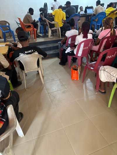 An indoor facility in Iriebe, Nigeria, displays a group of approximately 15-20 individuals in what appears to be a training or computer lab setting. The room features a light brown/beige tiled floor and plain white/off-white walls. The scene is divided into two levels: a lower ground area and a raised platform at the rear.

In the foreground and midground, on the lower level, several individuals are seated in rows of mixed-color plastic chairs (white, pink, red, light green). These chairs include specific markings like "025 SSHSS" and "007 SSHSS." One individual in the foreground, facing away from the camera, wears a black long-sleeve shirt with "MONEY" and dollar signs printed on the back, along with a black cap, seated in a white plastic chair. To their right, a person with reddish-brown braided hair is seated in a pink plastic chair. An orange shopping bag with white lining rests on the tiled floor between two chairs. Several individuals hold papers or documents.

On the raised platform at the back, approximately seven individuals, mostly adults, are seated at desks. At least five desktop computer monitors are visible, displaying what appear to be data or spreadsheet interfaces. A man in a bright yellow shirt is actively engaged with a computer. Another man in a white t-shirt stands behind a desk, near a standing electric fan. One person on the platform wears a shirt marked "BMSS 021." The individuals on the platform are operating computers, while those on the lower level are seated and observing or waiting.