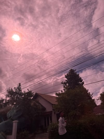 The image depicts an outdoor scene with a dusky, pink-hued sky filled with clouds. A bright, sun-like object is visible in the upper left quadrant, possibly lens flare or a partially obscured sun. Several power lines traverse the sky diagonally from the top left to the bottom right. In the middle ground, a house with a sloping roof is partially obscured by dense foliage. To the right of the house, a young person wearing a light-colored hooded jacket is standing and looking to their left. The foreground features a white pillar on the left and more greenery. The overall impression is a suburban or rural environment in Buengbakjok, Indonesia, at either dawn or dusk, characterized by an atmospheric sky and natural elements.