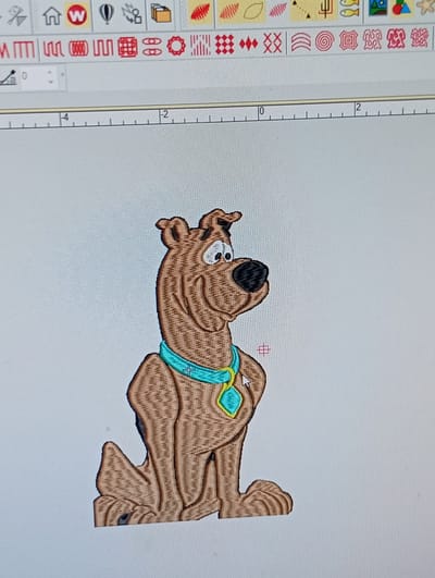 The image shows a computer screen displaying an embroidery design software. On the screen, a digitized image of the cartoon character Scooby-Doo is visible. The character is depicted in brown, with a blue collar and a green diamond-shaped tag. The software interface includes toolbars with various icons, a ruler with numerical markings, and a crosshair cursor. The background of the screen is a light gray. There is no information in the image to suggest any connection to Rumuepirikom, Nigeria.