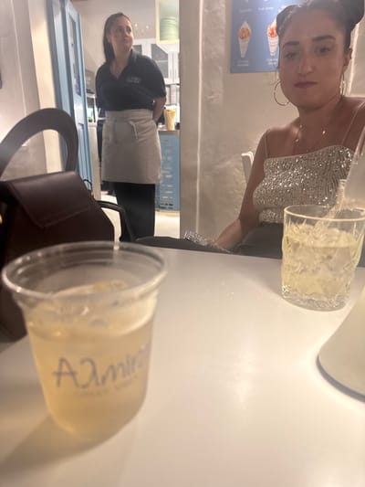 This media file depicts a woman seated at a white table, likely in a restaurant or cafe in Naousa, Greece. She is wearing a sparkly silver top and a delicate necklace. A glass of a pale yellow beverage, possibly a cocktail or lemonade, sits in front of her. Her hair is pulled back, and she has hoop earrings.

In the background, a person wearing a dark shirt with a logo and a light-colored apron is visible, suggesting they are likely an employee at the establishment. There's a glimpse of a blue door and shelves with what appear to be plates or serving dishes. The lighting suggests it is evening.

Another drink, in a clear plastic cup with a lid, is also on the table, showing blurry text that appears to read "Admir" or similar. The overall scene suggests a casual dining or social gathering.