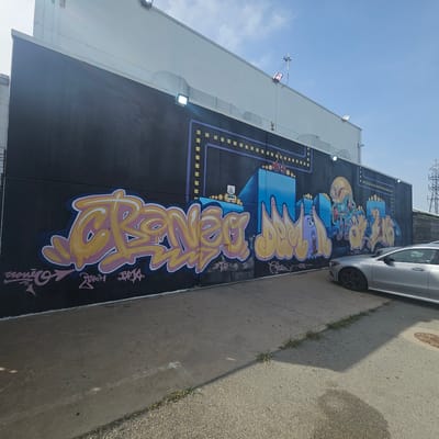 The image shows a mural on the side of a building. The mural is black with graffiti that is yellow and pink. The graffiti appears to be in a bubble letter style. The mural also has images of characters that appear to be from a video game. In front of the mural is a sidewalk and a silver car. The building is white and appears to be a commercial building. The sky is blue and there are a few clouds. The image is taken from a slightly low angle, looking up at the mural. There is no indication that this image is from the city of Girona, Spain.