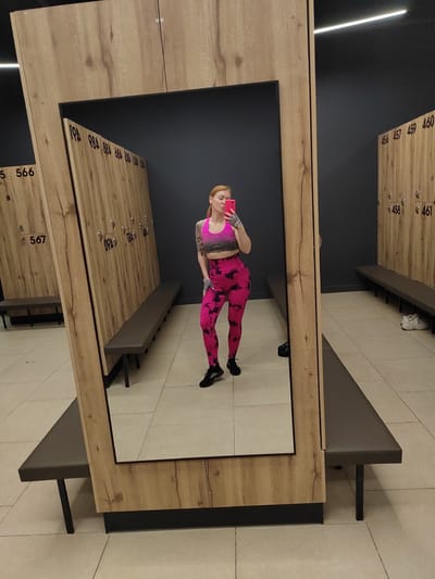 A woman is taking a selfie in a locker room in a building located in Novogorelovo, Russia. The woman has red hair and is wearing a pink and gray sports bra and pink leggings. She is standing in front of a large mirror that reflects her and the surrounding locker room. The locker room has wooden lockers with numbers on them, and benches are placed in front of the lockers. The floor is tiled. The lighting is bright, with overhead fluorescent lights visible in the reflection.