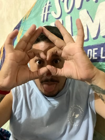 A medium close-up depicts an adult male from the chest up, centered in the frame. He has dark hair, a mustache, and a goatee. He wears a light blue sleeveless t-shirt with a circular white logo on the left chest. The logo text includes "BREEZE SURF COMPANY ESTABLISHED SIXTY NINE POINT BELLS BEACH QUALITY PROD." A red band is present on his left wrist, and tattoos are visible on both arms. The man's hands are positioned to form two circles or "okay" gestures directly in front of his wide-open eyes. His tongue is extended from his mouth. The background features a crumpled fabric or banner displaying large, light-colored lettering against a blue and turquoise pattern. The specific location within Bernal Este, Argentina, is not discernible from the visual evidence.