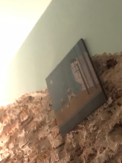 A rectangular framed print is affixed to a roughly textured, light-colored brick or stone wall. The print has a reflective surface showing a reflected interior space of what looks like a building. The wall is juxtaposed against a light blueish-green ceiling. Due to the lack of clear information, it is difficult to confirm the location in Beccar, Argentina. No people, actions, or interactions are visible.
