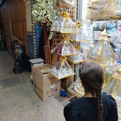 The image captures a scene in the city of Mechouar Fes Jdid, Morocco. A young girl with a long braid stands facing a display of gift baskets covered in clear plastic and adorned with golden ribbons. The baskets are arranged on shelves, showcasing various items within. To the left, a man is sitting on the ground with his head down, holding a dark bag. A bicycle leans against a wall behind him. Next to the man is a stack of boxes, one labeled "Induscap sa." A blue broom is visible behind the box. The background shows a narrow street with patterned tilework on the walls, typical of Moroccan architecture. The lighting suggests an indoor or covered market environment.