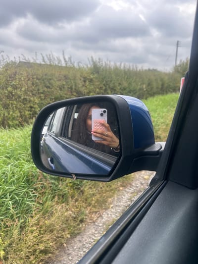 The image depicts a view from within a vehicle, focused on the driver's side wing mirror, with the setting identified as Winslow, United Kingdom. The mirror, featuring a blue housing and black frame, reflects a person with dark hair holding a light-colored smartphone with a pink, bubble-patterned case. A ring is visible on the reflected individual's right ring finger, which is grasping the phone.
The external environment visible through the window and around the mirror consists of dense green foliage, appearing as a hedgerow or overgrown bushes, on the left side of the frame. Beyond this vegetation, a distant building with a dark roof is partially visible. To the right of the mirror, tall green grass borders a dirt or gravel track. A utility pole and a partial red and white marker are visible in the far background. The sky is heavily overcast with grey clouds. Part of the vehicle's dark-colored interior door panel and window frame are visible in the foreground.