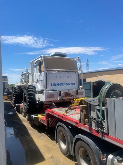 In Kungrad, Uzbekistan, a large, white, all-terrain vehicle with six massive wheels is being transported on a red flatbed truck. The vehicle has "БАШНЕФТЕГЕОФИЗИКА" written in blue Cyrillic letters on its side. The truck is parked on a dirt surface, with puddles of water visible, suggesting recent rain or a wet environment. In the background, there is a light-colored building with a blue-doored garage and a cell tower. A yellow forklift is partially visible to the left of the truck. The sky is bright blue with scattered white clouds.