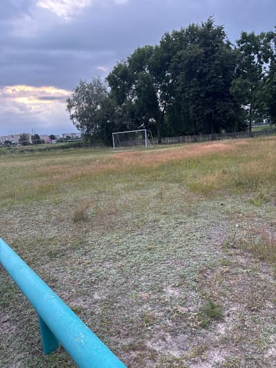 The image depicts an outdoor scene on a cloudy day. In the foreground, a bright turquoise-colored railing runs diagonally from the bottom left corner upwards. Beyond the railing is a field covered in dry, sparse grass with patches of lighter green vegetation and some small white flowers. In the middle ground, a white soccer goal stands in the center of the field. A weathered wooden fence runs horizontally behind the goal, separating the field from a line of mature trees with dark green foliage. In the background, a hazy view of buildings and structures suggests a populated area. The sky is filled with heavy, grey clouds, with a sliver of bright, pale yellow light breaking through on the left side, possibly indicating a sunset or sunrise. The environment appears to be a sports field or a park area on the outskirts of a town or village in the vicinity of Сароцкі сельскі Савет, Belarus. There are no people or animals visible in the image, and no specific actions or interactions are depicted. The scene is static and focuses on the landscape and a soccer goal.