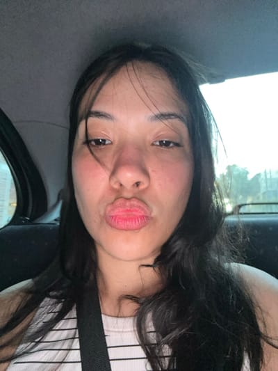 The image portrays a female individual seated within the interior of a vehicle in Villa Yapeyú, Argentina. Her long, dark hair cascades around her shoulders. She has a fair to medium complexion, with her dark eyes fixed on the camera. Her lips are pursed into a distinct 'kissing' expression, highlighted by a red color. She is attired in a light-colored top with horizontal stripes, and a black seatbelt diagonally crosses her chest. The vehicle's grey headliner is visible above. Through the vehicle's windows, the external environment is blurred, showing indistinct structures or light on the left and a lighter, possibly outdoor scene with foliage or sky on the right, consistent with ambient daylight.