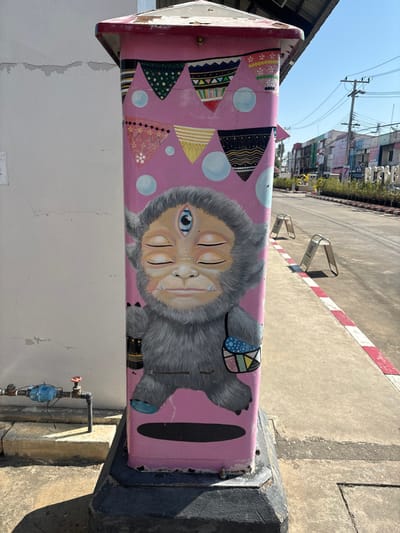 A pink mailbox, adorned with whimsical art, stands outdoors. The artwork includes colorful triangular pennants and a grey furry creature with four eyes, one open. The creature wears a patterned shoulder bag and wristband. The mailbox is mounted on a dark post. The background features a street scene with buildings and sidewalks, with a red-and-white striped curb. A water meter is visible nearby on the wall.