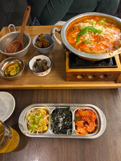 The image displays a meal set on a dark wooden table in a restaurant located in Chevilly-Larue, France. The upper portion of the frame features a light wooden serving tray. Centrally positioned on the tray is a round, silver-colored metal pot with two handles, containing a reddish-orange stew topped with melted white shredded cheese and chopped green scallions. This pot rests on a black metal trivet, which is supported by a wooden base with three circular cutouts on its visible side. To the left of the main pot, a smaller silver-colored metal bowl contains a wooden ladle. Adjacent to this, a small, light blue-grey ceramic bowl holds several dark brown, irregularly shaped fried items. Below these, a small silver-colored metal bowl contains sliced green cucumbers submerged in a translucent reddish-brown liquid. Another small, white ceramic bowl adorned with blue and red patterns contains dark, segmented items, consistent with pickled vegetables.
Directly on the dark wooden table, in the lower section of the image, is a silver-colored rectangular tray divided into three compartments. The left compartment holds light-colored bean sprouts garnished with green scallions. The middle compartment contains dark green, rectangular sheets of dried seaweed. The right compartment holds reddish-orange fermented cabbage, identified as kimchi. To the bottom-left, a clear glass filled with a yellowish liquid is partially visible. A portion of a white ceramic plate is also visible to the left of the three-compartment tray. In the upper background, a person wearing a dark green long-sleeved garment is partially discernible.