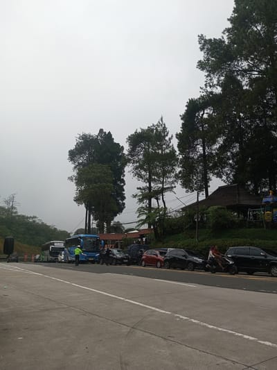 The image depicts a roadside scene on a cloudy day. A row of parked vehicles, including buses and cars, lines the side of the road. A traffic officer in a bright green vest stands near one of the buses. A motorcyclist with a passenger drives along the road next to the parked cars. Behind the vehicles, there is a grassy embankment with trees and a building with a traditional-style roof. The environment suggests a location outside of a dense urban area, likely in a rural or suburban setting. The location is in Mae Chan District, Thailand.