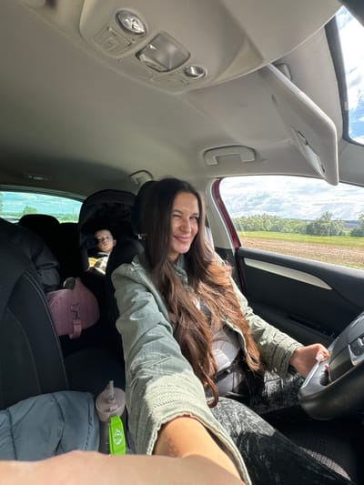 A woman sits in the driver's seat of a car, turned towards the camera, winking and smiling. She has long brown hair, wearing a light green jacket over a grey shirt. In the backseat is a baby in a car seat, a pink purse, a water bottle, and a grey puffy jacket. The landscape visible through the car windows suggests a rural area. The car's interior lights and sun visor are visible. The location is Kremyonki, Russia.