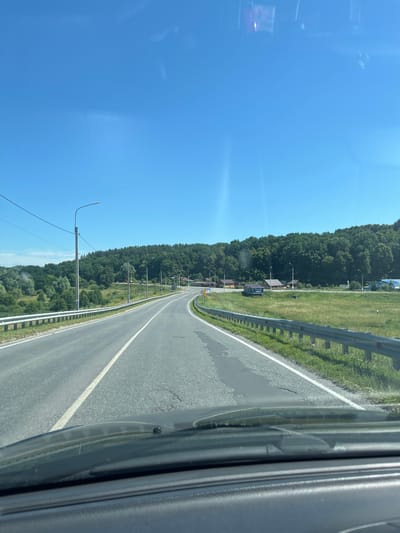 This is a vertical, eye-level, outdoor shot of a road in Yadrin, Russia, likely taken from inside a vehicle. The image is dominated by a bright blue sky on the left, with a reflective surface, possibly a window, distorting the view of the sky and some faint shapes within it.
The road, with a white dividing line, curves to the right and recedes into the distance. The asphalt appears worn in places. To the left of the road is a grassy embankment bordered by a metal guardrail. Beyond the embankment, a dense line of dark green trees forms a forest. Scattered amongst the trees, and some visible on the grassy area, are several small buildings with dark roofs, suggesting a village or settlement. Streetlights with curved tops are positioned along the side of the road, with power lines connecting them.
In the foreground, on the right side of the image, the interior of a vehicle is visible, with a dark, textured surface and reflections. A small black object is attached to this surface.
The scene appears to be set on a clear, sunny day, likely mid-morning or afternoon, given the bright sky and shadows. The overall mood is serene and calm, conveying a sense of travel through a rural landscape. One sign is visible on the side of the road, partially obscured, but the word "ПАРКА" (PARKA) can be read.