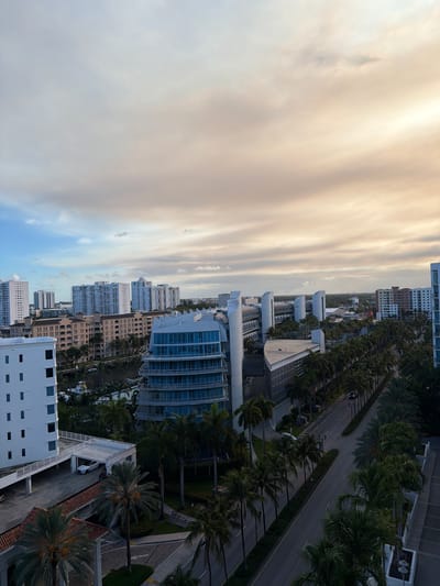 The image captures a cityscape in Aventura, Florida, USA, likely during the late afternoon or early evening, given the soft lighting and colorful sky. The scene is dominated by several modern buildings, including residential towers and commercial structures. Palm trees line the street, adding a tropical element to the urban environment.
In the foreground, a white multi-story building with numerous rectangular windows is visible on the left. Adjacent to it is a covered parking structure with a white roof, where a white car is parked. A road curves through the scene, bordered by a row of palm trees on either side. In the middle ground, a distinctive curved blue and white building stands out, reflecting the sky. Beyond it, a cluster of taller residential buildings, mostly white, stretches across the horizon.
In the distance, more buildings and urban development are visible, fading into a hazy sky. The sky itself is a prominent feature, with soft hues of blue, white, and orange clouds, suggesting either sunrise or sunset. A few boats can be seen in a waterway in the middle left of the image, indicating proximity to water. There are no people clearly visible in the image. The overall impression is one of a well-developed, modern urban area with a tropical atmosphere.