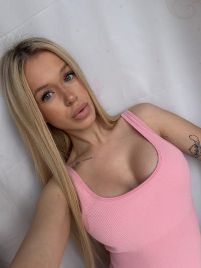 The image shows a top-down, close-up shot of a young woman lying down on a white surface with a subtle pink dot pattern. She has long, blonde hair and is wearing a light pink ribbed top. Her face is turned towards the camera, with her eyes open and looking forward. She has visible makeup, including mascara and lip gloss. A small nose ring is visible on her septum, and she has a tattoo of a cartoon character on her upper left arm. There is also a tattoo on her neck with some illegible text. The lighting suggests it is daytime, and the setting appears to be indoors, possibly a bedroom or studio, given the smooth white surface and the subtle decorative pattern. The overall mood is relaxed and intimate.