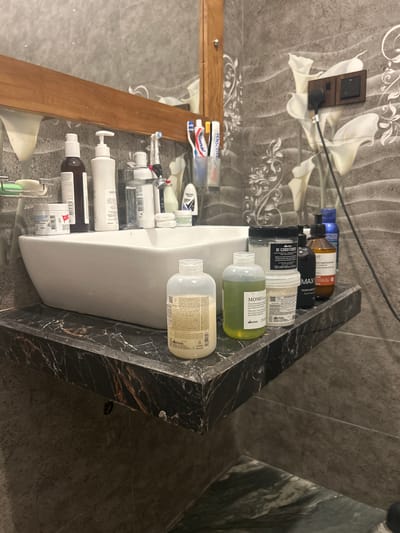 The image shows a bathroom vanity counter with a white vessel sink. Various toiletries are arranged on the counter, including bottles of shampoo, conditioner, and lotions, as well as toothbrushes. The counter is made of black marble with prominent white veining. The wall behind the sink is tiled with a gray, textured pattern and features a decorative floral design. A wooden-framed mirror is mounted above the sink. A dark-colored soap dispenser and a bottle of blue liquid are also visible on the right side of the counter. A power outlet with two sockets is mounted on the wall to the right of the mirror. The overall impression is that of a clean and organized bathroom. The location is identified as Kadewatta, Sri Lanka.