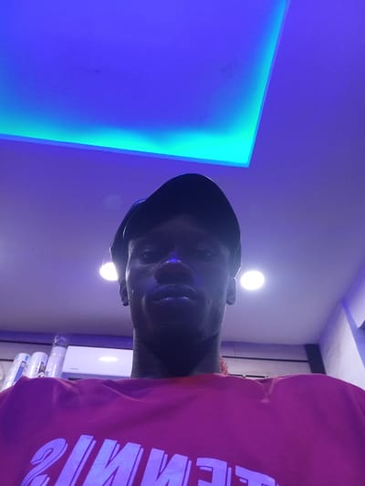 A selfie-style, low-angle portrait of a dark-skinned man wearing a black cap and a pink t-shirt with white lettering is set in an indoor environment. Two round light fixtures are visible on the ceiling, and an LED panel emits a blue glow. White containers and an air conditioning unit are visible in the background. The location appears to be in Utagban, Nigeria.