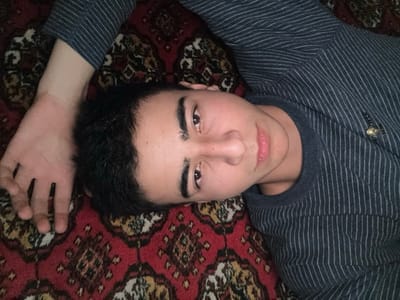 A young man lies on a patterned rug, facing the camera. He is wearing a striped, long-sleeved shirt. His left arm is extended above his head, the hand near the top edge of the frame. He has dark hair, prominent eyebrows, and is looking directly at the viewer with a slight smile. The rug has a complex, traditional design, primarily in red, brown, and gold.