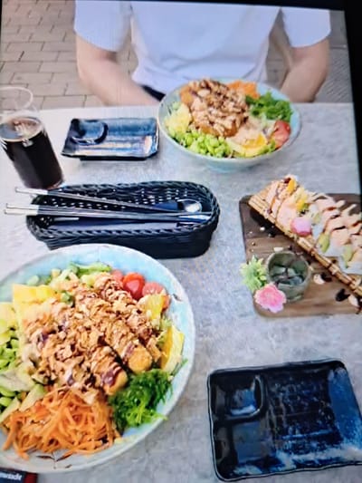 A high-angle shot displays an array of food items arranged on a surface, seemingly a table, with a person seated at the left edge. On the left, a person wearing a white t-shirt is partially visible. Two bowls of food are presented, each containing a mix of fresh ingredients such as vegetables, leafy greens, and toppings. A wooden serving board is visible, showcasing what appears to be sushi rolls, arranged with decorative elements. Dark-colored plates are placed around the table. In the center, there is a holder containing eating utensils. A dark can is situated near the utensil holder. The surroundings and the setting's lighting suggest an indoor location. There is no indication in the image that it is in Wuppertal, Germany.