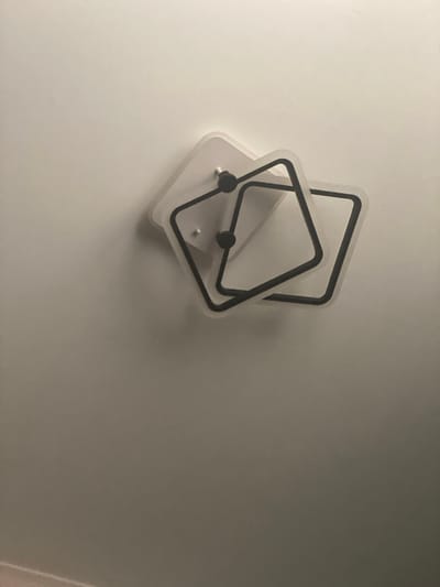 This media file features two interlocking, clear plastic frames with black borders, mounted on a white, textured wall. Each frame is square-shaped but rotated, creating an overlapping diamond pattern. Two small, dark circular objects, possibly fasteners or decorative elements, are visible on the lower frame, one slightly above and to the left of the other. The lighting suggests an indoor setting, possibly a room, with the light source casting a soft glow on the wall and illuminating the translucent frames. The arrangement of the frames appears static, with no apparent activity taking place. The background is a plain white wall, providing a neutral backdrop for the decorative object. No text is visible in the image. The overall impression is one of minimalist, modern decor.