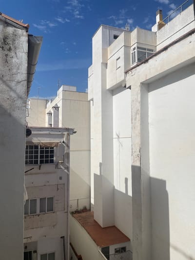 This is a photograph taken from a low angle looking upwards at a series of white buildings against a bright blue sky with scattered white clouds. The buildings appear to be residential or commercial structures, with multiple floors and visible windows. Some of the buildings have balconies with railings. There are also some architectural details visible, such as concrete beams and ledges, some of which show signs of wear and tear, including cracks.
In the foreground, there is a textured, weathered wall in white and grey tones, possibly a rooftop or terrace parapet. A dark, cylindrical object, possibly a pipe or conduit, runs along the top of this wall and then curves upwards.
Further back, a darker, curved structure is visible on the roof of a lower building, with visible metal bands. Antennas and wires are also present on the roofs of some of the buildings, suggesting they are inhabited or used for communication.
The light suggests it is daytime, likely sunny due to the strong shadows cast by the buildings. The sky indicates clear weather. The architecture and the presence of overhead wires are common in urban or semi-urban Mediterranean environments. The scene appears to be a snapshot of a typical urban landscape in Sagunto, Spain. No people are visible in the image.