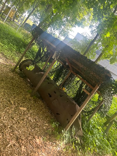 The image shows an outdoor scene with a large, rusted, cylindrical object lying on its side, partially covered by a wooden structure with camouflage netting. The object appears to be a piece of industrial pipe or a large artillery shell. The ground is covered with wood chips, and lush green vegetation surrounds the structure and the object. Trees with dense green foliage create a canopy overhead, with sunlight filtering through the leaves. In the background, a modern building with white walls is visible through the trees. The scene is likely part of an outdoor exhibit or a historical display. The location is identified as being in Shlisselburg, Russia. There are no people visible in the image.