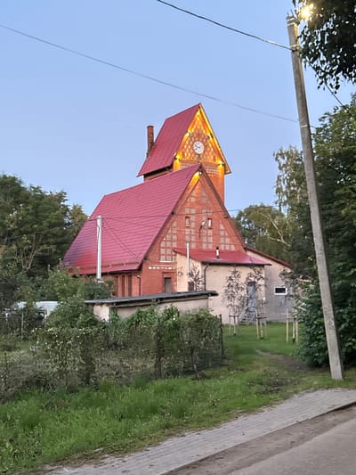 The image depicts a brick building with a red roof, likely a church or a similar public structure, with a clock face visible near the apex of the roof. The building is surrounded by greenery, including trees and a lawn. To the right of the building, a paved walkway and a road are visible. A utility pole with wires runs across the top left portion of the image, with a light fixture attached. The sky is clear and blue, suggesting it is daytime. The overall scene appears to be a quiet, possibly rural or suburban setting in Primorye, Russia. There is no visible text in the image.