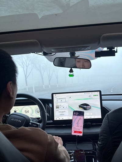 The scene depicts the interior of an AION brand vehicle in motion, viewed from a rear passenger's perspective looking forward. An adult driver, visible from behind with dark, short hair, is positioned at the black steering wheel, which features the "AION" logo. The digital instrument cluster behind the steering wheel shows a speed of 54 km/h. A large, horizontal infotainment screen on the center dashboard displays an active navigation map featuring a 3D vehicle model and a music playback interface. Below this main screen, a smartphone is vertically mounted, exhibiting a navigation application (likely Gaode Maps) with a reported remaining distance of 172 km.
A small, green, droplet-shaped air freshener hangs from the rearview mirror, which reflects the upper portion of a passenger's face, likely an adult female. A black dashcam device is affixed to the windshield to the right of the rearview mirror. The external environment, viewed through the windshield, is characterized by dense fog or haze, severely limiting visibility. Faint outlines of bare trees and white road barriers are discernible along a paved roadway. The vehicle is actively navigating within Huangdao District, China, under these low-visibility conditions.