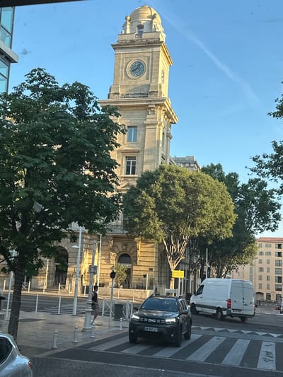 The image depicts a street scene in Toulon, France, featuring a tall, stone clock tower with a dome as the central architectural element. The tower has a visible clock face. In the foreground, a dark-colored SUV is stopped at a pedestrian crossing, with its license plate reading "GM-587-LF". To its right, a white cargo van is in motion, heading away from the camera. A pedestrian wearing a dark tank top and shorts is walking near a lamppost on the sidewalk. Several trees with lush green foliage line the street, some casting shadows. The background includes a multi-story building with a classical facade and windows, and further in the distance, another building with a distinctive tiled roof. The sky is clear blue with a faint contrail. The overall scene suggests an urban environment during daylight hours.