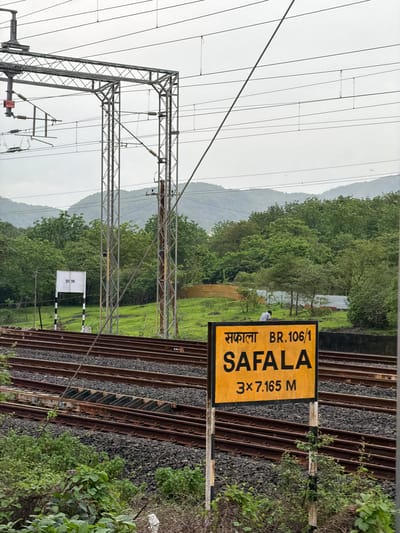 This is a medium shot of a train station sign in Saphale, India, on a cloudy day. The sign, which is yellow with black lettering, reads "Safala BR.106/1" and "SAFALA 3x7.165 M". It is mounted on a pole with black and white stripes. In the background, there are train tracks, overhead electrical wires, and lush green trees on a hillside. A few birds are visible in the sky. The scene appears to be at an outdoor railway station.