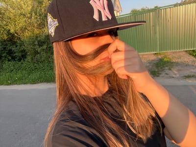 This media file is a close-up, medium shot of a person wearing a black baseball cap with pink lettering. The cap has a logo patch on the side. The person is holding a section of their long, brown hair in front of their face, obscuring their mouth and nose. Only one eye is fully visible. The person is wearing a black t-shirt. The scene appears to be outdoors during the golden hour, as evidenced by the warm, directional lighting casting shadows. The background includes lush green foliage on the left and a tall, ribbed green metal fence on the right. A paved surface, likely a road or pathway, is visible in the foreground. The overall impression is casual and playful, with the person seemingly posing for a selfie. The time of day is late afternoon or early evening. The location context provided is Pushkino, Russia. There is no discernible text other than the lettering on the baseball cap.