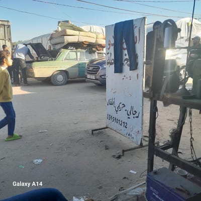 The image shows a street scene in Al-Mawasi, location unknown. In the foreground, there's a sign in Arabic with a pair of pants hanging on it, possibly advertising clothing. A person wearing a yellow shirt, blue pants, and green shoes is lying on the ground near the sign. Behind them, two other people are standing next to an older green van with items strapped to its roof. A modern blue car is parked behind the van. The background suggests a cluttered urban environment.