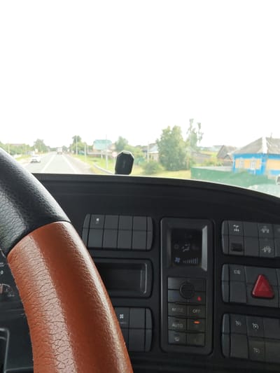 The image shows the interior of a vehicle's cabin, looking forward through the windshield. A portion of the steering wheel, covered in black and brown material, is visible on the left. The dashboard features several buttons and a digital display, likely for climate control or vehicle information. The background shows a rural landscape with a road, trees, and houses, suggesting the vehicle is on the road in a village. The houses appear to be of traditional Russian architecture, with brightly colored elements. The sky is overcast, indicating a cloudy day. The location is specified as Новые Выселки, Russia. There are no people visible in the frame. The scene is static, depicting a moment in time while the vehicle is in transit.