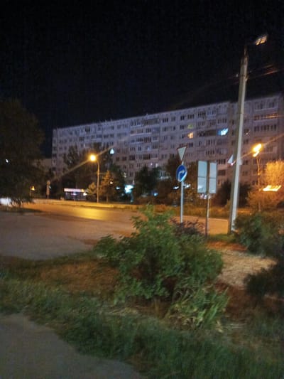 The image depicts a nighttime street scene in Volzhsky, Russia, featuring a large, multi-story apartment building as the dominant background element. The building appears to be constructed of concrete panels and has numerous windows, many of which are illuminated, suggesting occupancy. In the foreground, a paved road curves to the left, with a grassy verge and dense shrubbery bordering its right side. A streetlamp casts a bright yellow light onto the road. Several traffic signs are visible: a triangular sign with a red border, a circular blue sign with a white arrow indicating a left turn, and two rectangular signs mounted on a pole. A utility pole with wires runs vertically along the right side of the frame, illuminated by streetlights. The overall impression is that of a residential area at night, with the city lights creating a somewhat hazy atmosphere.