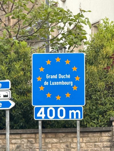 A road sign indicating the Grand Duchy of Luxembourg is 400 meters ahead, situated in an outdoor setting with vegetation and a stone wall nearby. The sign is blue with yellow stars and white lettering. Other blue road signs are visible. The setting appears to be Aubange, Belgium, as the sign suggests proximity to Luxembourg. No people are visible.